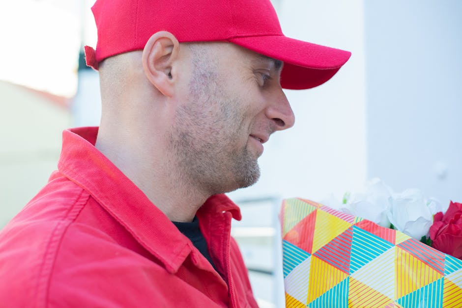 A man wearing a red cap and red jacket is holding a colourful floral gift bag filled with fresh flowers, including red and white blooms. He is smiling softly while looking at the flowers, which are ar