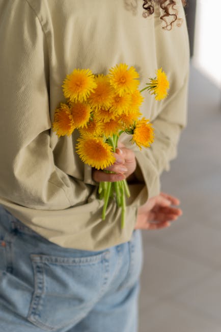 A person in a beige long-sleeve top and light blue jeans is holding a bouquet of vibrant yellow dahlias with full, layered petals and green stems. The bouquet is gathered in one hand, with some flower