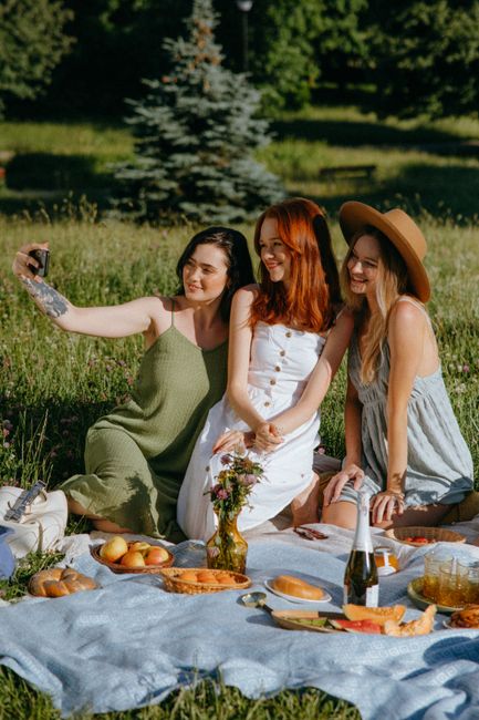 Three young women with cheerful expressions are enjoying a picnic on a lush green lawn near Bushy Park, with trees and sunlight filtering through the leaves in the background. They are sitting on a li