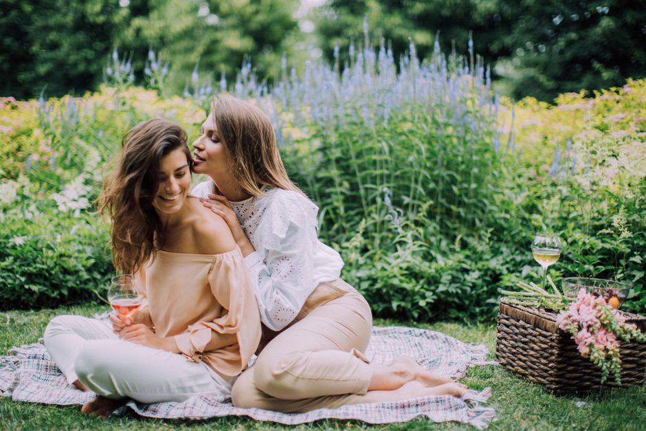 Two women sitting on a checkered picnic blanket in a lush garden with vibrant green foliage and flowering plants in the background. One woman with long, wavy brown hair, wearing a beige off-the-should