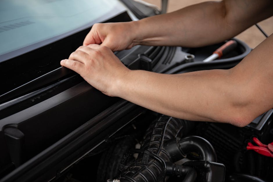 A person’s hands carefully inspecting and adjusting the engine compartment of a vehicle, with visible hoses, wires, and engine components beneath the open hood, in a well-lit setting. This image is