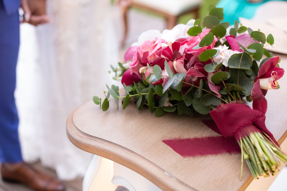 A vibrant bridal bouquet featuring pink and white orchids, complemented by lush green eucalyptus leaves, rests on a light wooden table decorated with a deep red ribbon tied around the stems. The bouqu