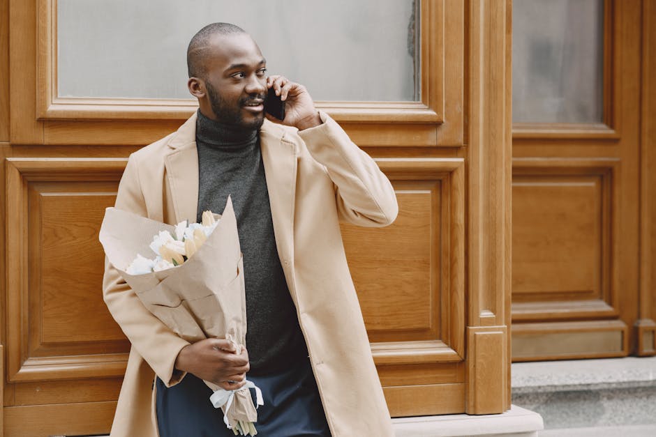 A man with dark skin and a neatly trimmed beard stands outside in front of a wooden panelled building, holding a bouquet of fresh white roses wrapped in brown craft paper. He is dressed in a beige tre